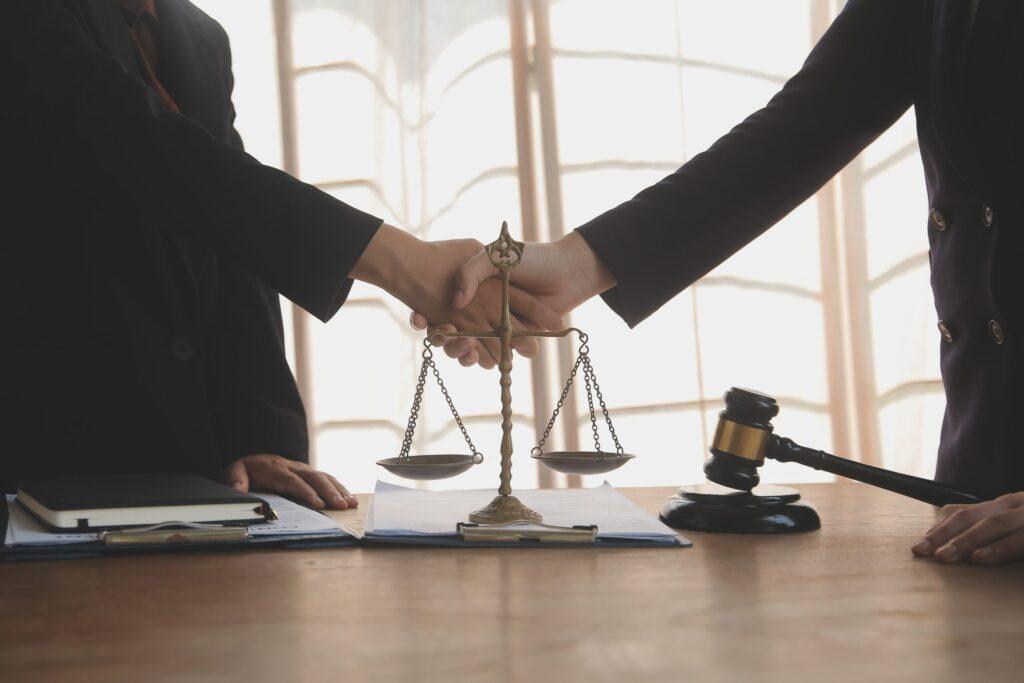 Lawyers shaking hands over contract with scales of justice and judge gavel on desk in law office.