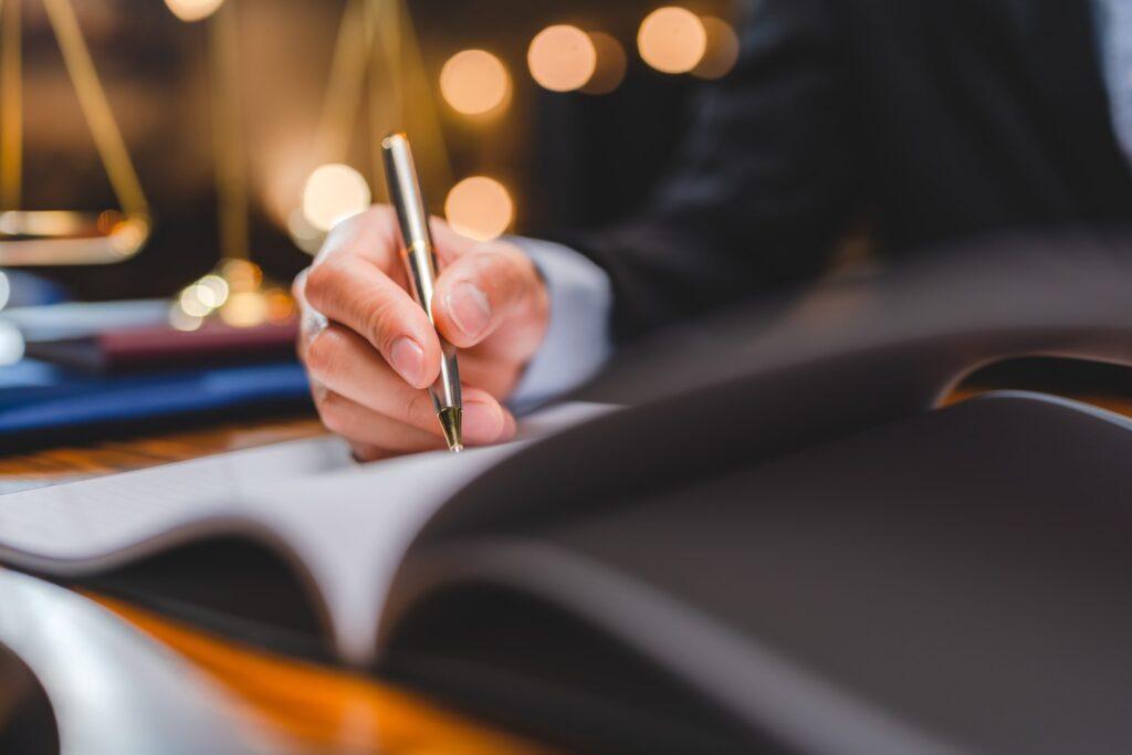 Close-up of lawyer writing notes in legal document with scales of justice in background, representing legal work or case preparation