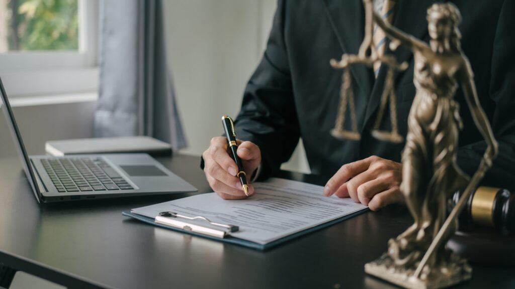 Attorney signing legal documents at office desk with laptop and Lady Justice statue in foreground.