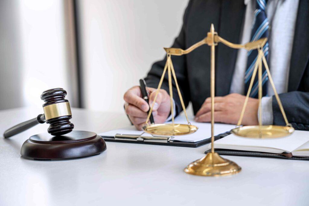 Lawyer writing notes at a desk with scales of justice and a judge&rsquo;s gavel, representing legal consultation, justice, and professional law services.