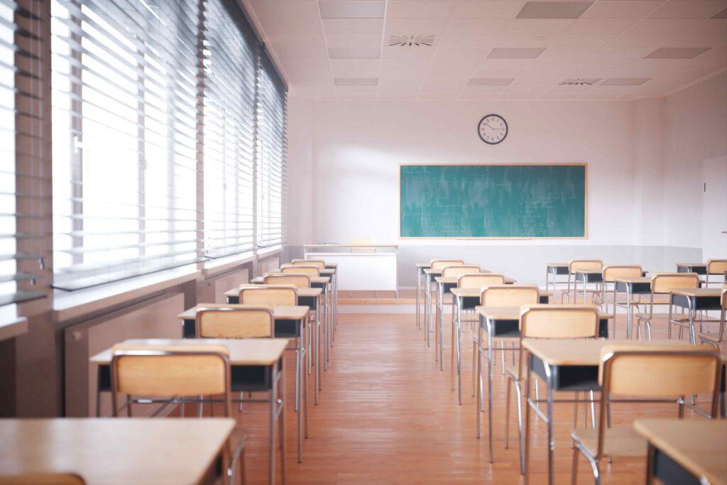 Empty classroom with rows of desks and chairs facing chalkboard, natural light from windows and school clock on wall